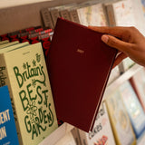 A hand holding a dark red hardcover planner or journal labeled '2027' against a blurred bookshelf background with other books visible.