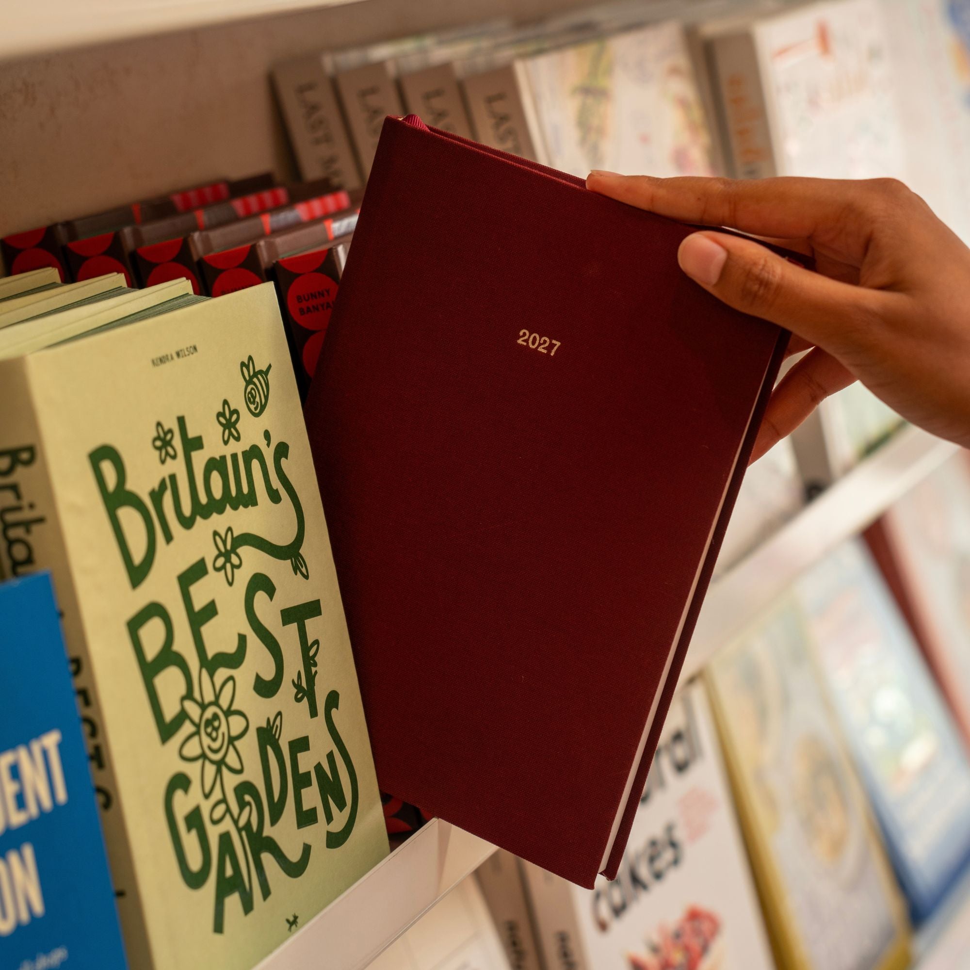 A hand holding a dark red hardcover planner or journal labeled '2027' against a blurred bookshelf background with other books visible.