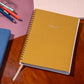 Yellow spiral-bound planner on a wooden surface with pens and another book in the background.
