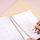 Person holding a pencil over a planner with a grid layout on a light pink surface