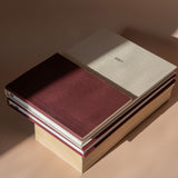 A stack of hardcover planners or notebooks on a warm beige surface, with one dark burgundy cover on top, a light beige cover labeled 2027 behind it, and additional stacked volumes underneath in cream and burgundy tones.