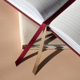 A close-up of an open notebook or journal with two ribbon bookmarks: one dark red and one metallic gold, resting on a beige fabric-covered book or box against a warm beige background.