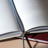 An open lined notebook or journal with a blue ribbon bookmark draped over the pages, resting on a beige surface with a dark red textured book underneath.