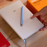A close-up of a person holding a beige 2027 planner or notebook with a light blue pen on top, placed on a wooden desk with other colorful books in the background.