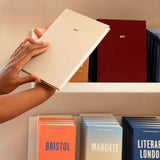 A person's hands holding a light beige 2027 edition of Papersmiths 2027 diary against a beige bookshelf, with other colorful guidebooks in the background.