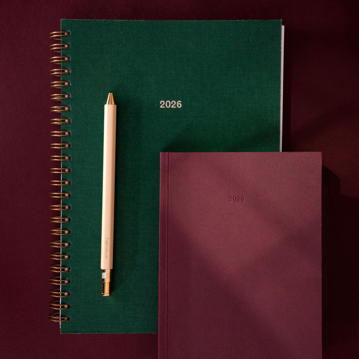 Two notebooks, one green and one burgundy, with a pen on a dark red background.