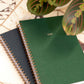 Green spiral-bound planner with '2026' on a marble surface with a plant.