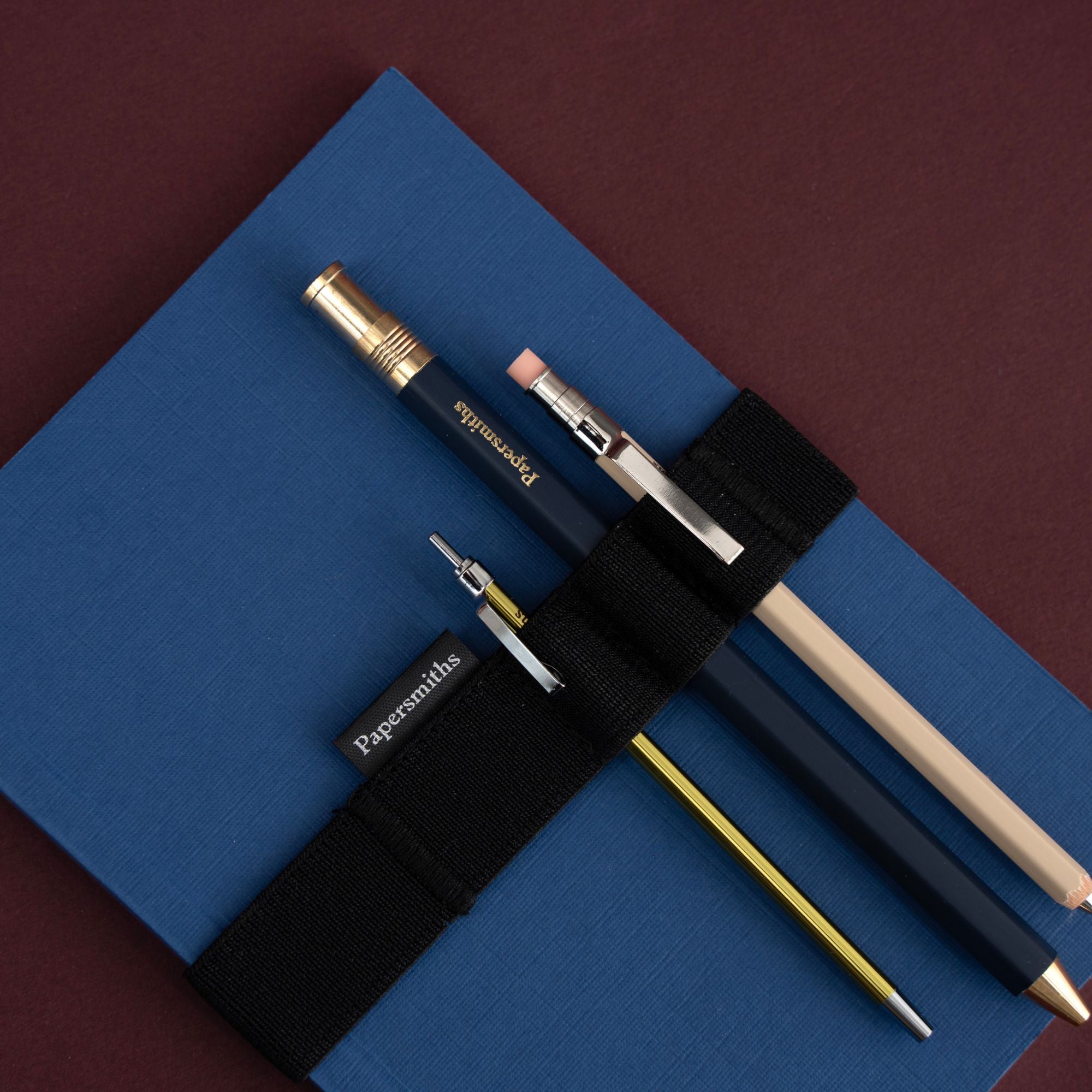 A set of three Papermiths pens and pencils secured in a black elastic loop over a blue notebook, set against a dark maroon background.