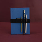 A blue softcover notebook with a black elastic band holding three pens, photographed against a dark burgundy background. The brand name 'Papersmiths' is visible on the pen labels.