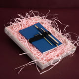 A blue notebook with a black strap and two pens, presented in a white gift box filled with pink shredded paper on a dark burgundy background.