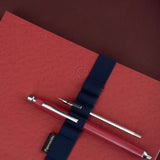 A red textured notebook with a navy blue elastic strap holding a red pen with silver metal accents. The brand name 'Papersmiths' is visible on a small black tag attached to the strap.