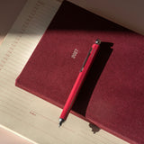 A red penwith silver accents rests diagonally on top of a textured burgundy fabric cover, which is placed on an open cream-colored lined notebook under warm, direct sunlight.