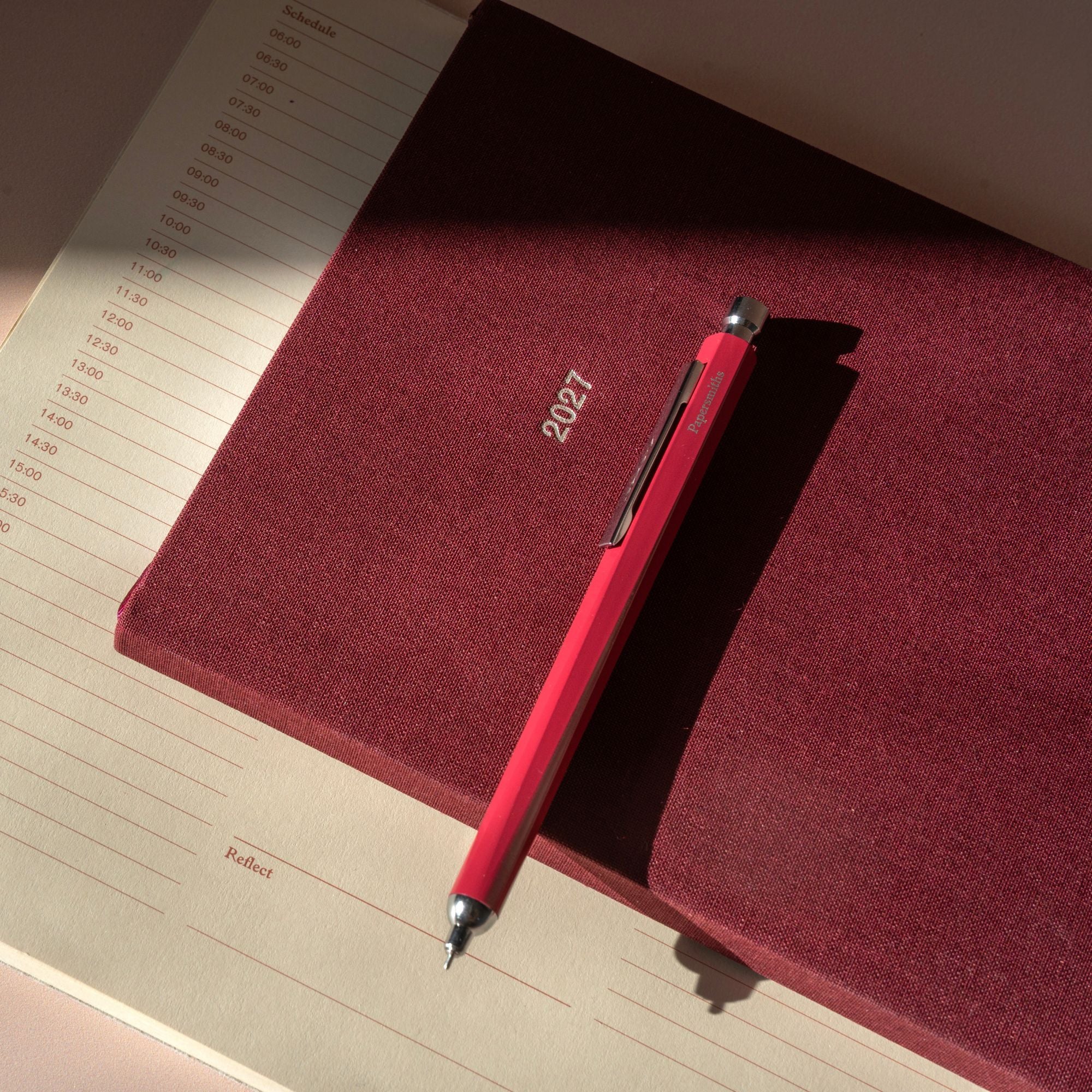 A red penwith silver accents rests diagonally on top of a textured burgundy fabric cover, which is placed on an open cream-colored lined notebook under warm, direct sunlight.