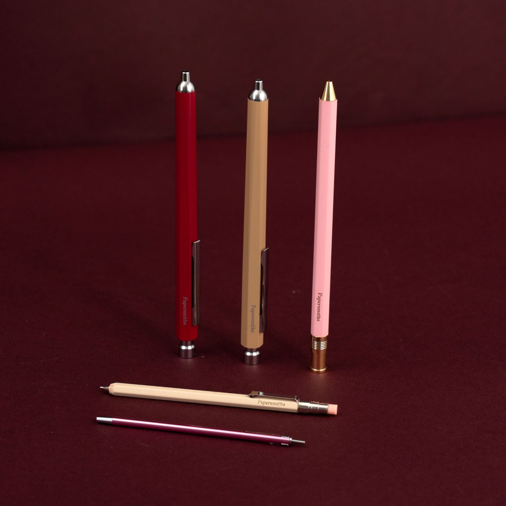 Three slim retractable ballpoint pens in red, tan, and pink are displayed upright on a dark burgundy background, with one tan pencil and a pink pocket pen lying horizontally in front.