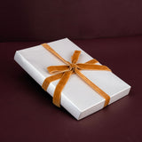 A white rectangular gift box wrapped with a wide tan ribbon tied in a bow, photographed on a dark burgundy background.